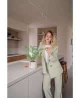Een vrouw met krullend blond haar in een licht pak staat in een moderne keuken en glimlacht terwijl ze zachtjes de bladeren van de Kunstplant Varen 35 cm potplant op het aanrecht aanraakt.