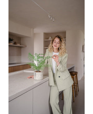 Een vrouw met krullend blond haar in een licht pak staat in een moderne keuken en glimlacht terwijl ze zachtjes de bladeren van de Kunstplant Varen 35 cm potplant op het aanrecht aanraakt.