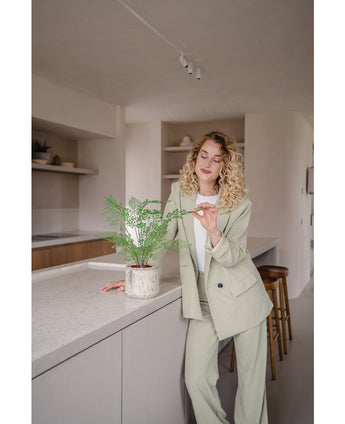 Een vrouw met krullend blond haar in een licht pak staat in een moderne keuken en glimlacht terwijl ze zachtjes de bladeren van de Kunstplant Varen 35 cm potplant op het aanrecht aanraakt.