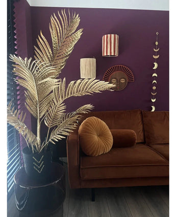 Een moderne woonkamer met een bruin fluwelen bank, rond plooikussen en de Kunstpalm Areca goud 120 cm in een zwarte vaas als gouddecor. Geometrische wandkunst hangt op een donkerpaarse muur, met houten vloeren en verticale lamellen in het zicht.