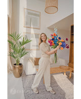 Een glimlachende vrouw in lichtbeige kleding en sneakers staat in een lichte, moderne woonkamer met houten meubels en ingelijste kunst, en houdt een kleurrijk boeket vast. De Kunstpalm Kentia 135 cm voegt een tropisch tintje toe aan de ruimte.