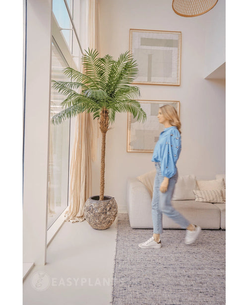 Une femme vêtue d'un chemisier bleu et d'un jean passe devant le Kunstpalm Phoenix De Luxe 210 cm près d'une grande fenêtre dans un salon lumineux et moderne avec des murs beiges, un canapé et des œuvres d'art encadrées au mur.