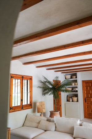 Een gezellige woonkamer met witte muren, houten balken, een witte sectionele bank, de Kunstpalm Phoenix De Luxe 210 cm, houten raam en deur, ingebouwde planken met manden en aardewerk, en natuurlijk licht dat door het raam naar binnen valt.