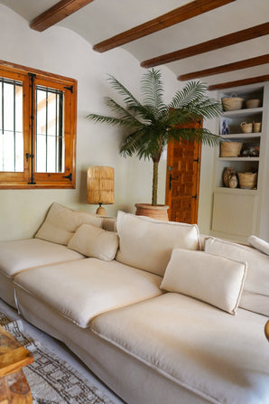 Een gezellige woonkamer met een grote crèmekleurige bank, de Kunstpalm Phoenix De Luxe 210 cm in de hoek, een houten lamp, ingebouwde planken met manden, houten plafondbalken en een raam met houten omlijsting.