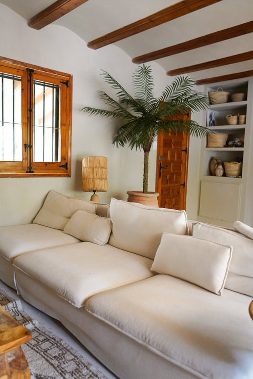 Un salon cosy avec un grand canapé couleur crème, le Palmier Artificiel Phoenix De Luxe 210 cm dans le coin, une lampe en bois, des étagères intégrées avec paniers, des poutres en bois au plafond et une fenêtre avec des cadres en bois.