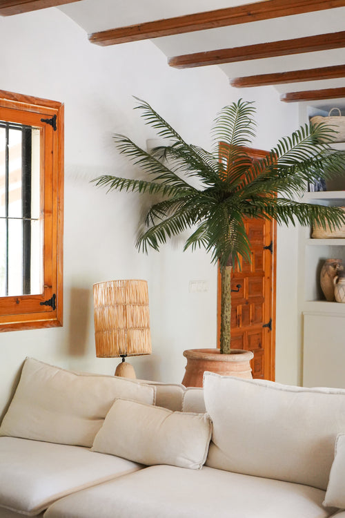 Un salon cosy avec un canapé blanc, une lampe de table en rotin, le Palmier Artificiel Phoenix De Luxe 210 cm dans un pot, des poutres en bois au plafond, une fenêtre avec des cadres en bois et une porte en bois en arrière-plan.