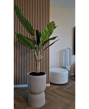 De Kunstplant Bananenboom 150 cm, met zijn weelderige groene bladeren, staat op een houten vloer bij een geribbelde wand met houten panelen. Vlakbij staan een witte ronde voetenbank en een ventilator zonder bladen op de achtergrond.