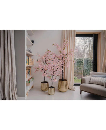 Een Kunstplant Bougainvillea 120 cm roze staat bij het raam in een gezellige woonkamer met witte muren, een boekenplank, beige gordijnen en een grijze fauteuil.