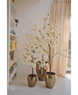 Een Kunstplant Bougainvillea 120 cm groot met witte bloesems in een goudkleurige pot met textuur staat bij een raam met transparante gordijnen, naast boekenplanken vol boeken en decor in een lichte, moderne kamer.