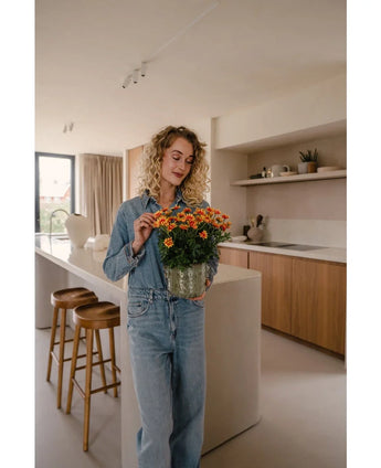 Een vrouw met krullend blond haar, gekleed in een denim shirt en spijkerbroek, staat in een moderne keuken met de Kunstplant Daisy UV 31 cm in haar hand en glimlacht er zachtjes naar.