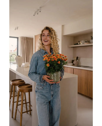 Een vrouw met krullend blond haar, in een denim shirt en spijkerbroek, staat in een moderne keuken met de Kunstplant Daisy UV 31 cm in haar hand, glimlacht en kijkt een beetje opzij.