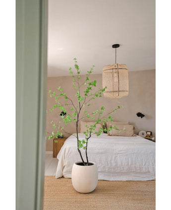 Een minimalistische slaapkamer met de Kunstplant Enkianthus 180 cm als grote potplant, een bed met wit beddengoed, een geweven hanglamp, beige muren en een vloerkleed van natuurvezel voor een eenvoudige en luchtige sfeer.