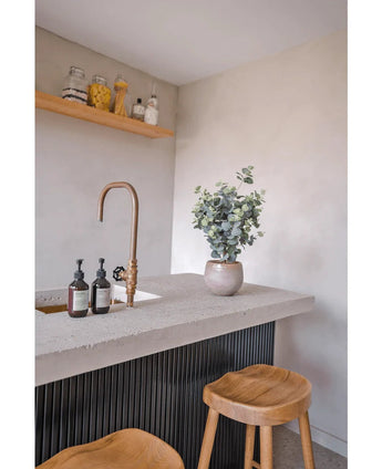 Een modern aanrecht heeft een messing kraan, zeepdispensers, de Kunstplant Eucalyptus 61 cm potplant en twee houten krukjes. Boven het aanrecht houden planken op een lichte muur potten en flessen.