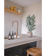 Een modern aanrecht heeft een gouden kraan, drie zeepdispensers, de Kunstplant Eucalyptus 91 cm en houten krukjes. Minimalistische planken boven tonen potten met verschillende ingrediënten voor een gezellige uitstraling.