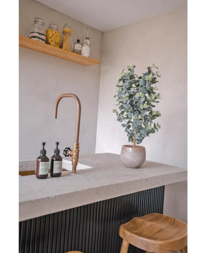 Een modern aanrecht heeft een gouden kraan, drie zeepdispensers, de Kunstplant Eucalyptus 91 cm en houten krukjes. Minimalistische planken boven tonen potten met verschillende ingrediënten voor een gezellige uitstraling.