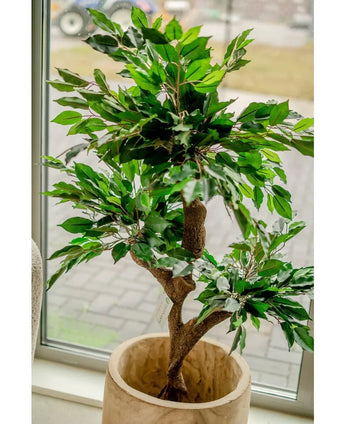 De Kunstplant Ficus Crazy 120 cm, met zijn dichte groene bladeren in een pot met beige structuur, staat bij een groot raam dat de kamer vult met natuurlijk licht. Stijlvolle sierpotten en een betegeld gebied zijn buiten zichtbaar door het raam.