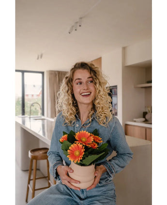 Een glimlachende blonde vrouw in spijkerstof zit in een moderne keuken met de Kunstplant Gerbera UV 28 cm oranje in de hand - een levendige kunstmatige oranje gerberapot. Zonlicht stroomt door een groot raam en verlicht de ruimte.