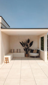 Een moderne, minimalistische patio met crèmekleurige tegels, een ingebouwde bank met gestreepte kussens, een Kunstplant Heliconia 210 cm voor een luxe touch, een klein houten krukje en een ronde stoel met kussens onder een plat dak tegen een helderblauwe hemel.