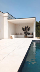 Modern buitenzwembad met lichte tegelvloer, een witte overdekte zithoek met gestreepte kussens, een Kunstplant Heliconia 210 cm voor een luxe touch, een klein krukje, een sofa, blauwe lucht erboven en een groene haag om het plaatje compleet te maken.