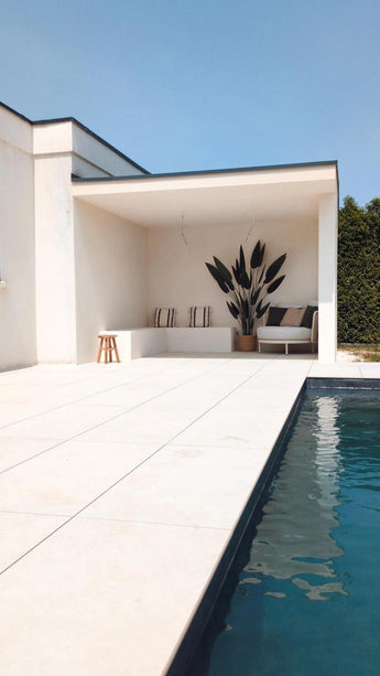 Modern buitenzwembad met lichte tegelvloer, een witte overdekte zithoek met gestreepte kussens, een Kunstplant Heliconia 210 cm voor een luxe touch, een klein krukje, een sofa, blauwe lucht erboven en een groene haag om het plaatje compleet te maken.