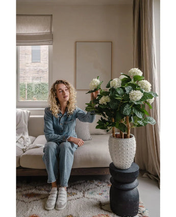 Een vrouw met krullend blond haar, in een spijkerpak en witte sneakers, zit binnen op een beige sofa en raakt voorzichtig de Kunstplant Hortensia UV 85 cm aan - een grote witte kunsthortensia in pot - die op een zwarte sokkeltafel staat.