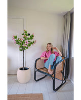 Une femme vêtue d'un pull rose et d'un jean bleu est assise pieds nus sur une chaise en rotin à côté de la plante artificielle Judas 150 cm, un arbre artificiel à fleurs roses, dans une pièce lumineuse et moderne aux murs et au sol blancs.