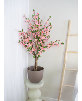 De Kunstplant Kersenbloesem 120 cm roze, een hoge kunstkersenbloesem met roze bloemen, staat op een witte ronde sokkel bij een wit gordijn en een lichte houten ladder tegen een lichte muur.