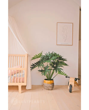 Een gezellige kinderkamer met een houten wieg, een Kunstplant Philodendron 90 cm in een grote pot, minimalistische lijnkunst aan de muur en een kleine houten loopfiets met het label "MIA" op een lichthouten vloer.