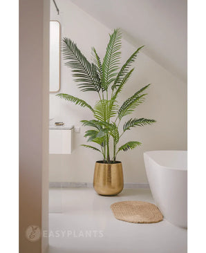 De Kunstplant Phoenix 180 cm in een gouden pot staat bij een wit bad en wastafel in een moderne minimalistische badkamer met witte muren en vloer. Vlakbij ligt een ronde geweven mat op de vloer.