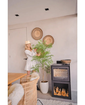Een vrouw in een witte jurk en hoed staat naast de Kunstplant Ruscus 150 cm, die een natuurlijke flair toevoegt aan een moderne kamer met rieten meubels, muurmanden en een zwarte houtkachel - perfect voor decoratieve interieurs.