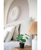 Een keurig opgemaakt bed met witte kussens en beddengoed staat naast een houten nachtkastje met de Kunstplant Watermeloen 29 cm en een lamp met een geweven kap; boven het hoofdeinde hangt een wandsculptuur met witte textuur.