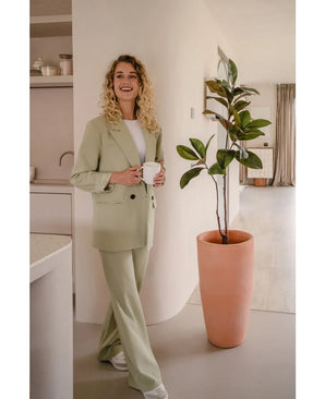 Een glimlachende vrouw met krullend blond haar, gekleed in een lichtgroen pak en witte sneakers, staat binnen met een witte mok naast de Kunstplant Yulaniaboom 120 cm in een moderne, neutraal gekleurde kamer.