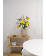 Een beige vaas met Kunstranonkel Real Touch Geel 60cm bloemen staat op een lichte houten plank naast een ronde witte tafel. Op de plank staan ook een kleine witte klok, een houten dienblad en een boek tegen een effen witte muur.