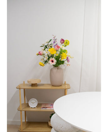 Een beige vaas met Kunstranonkel Real Touch Geel 60cm bloemen staat op een lichte houten plank naast een ronde witte tafel. Op de plank staan ook een kleine witte klok, een houten dienblad en een boek tegen een effen witte muur.