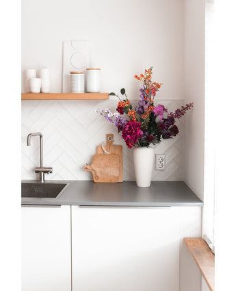 Une cuisine blanche moderne avec un plan de travail gris, un évier, une planche à découper en bois et un vase blanc rempli d'hortensia artificiel de 55 cm rose foncé. Des tasses et des pots blancs sont posés sur une étagère en bois au-dessus du comptoir.