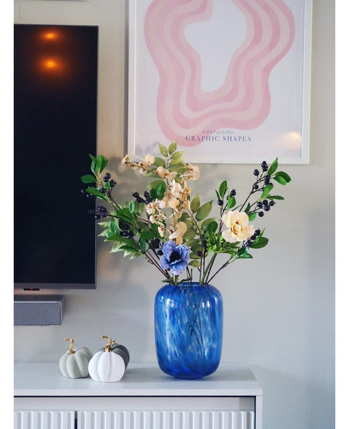 Un vase en verre bleu avec une fleur artificielle violette de 76 cm de branche artificielle se dresse sur une armoire blanche. À côté se trouvent deux citrouilles blanches aux tiges dorées. Sur le mur derrière, sont accrochés une télévision et une reproduction d'art abstraite rose.