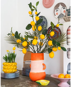 Een lichte kamer met een hoge vaas vol citroenen en groene bladeren, waarin de opvallende Kunsttak Lemon 104 cm geel een prominente plaats inneemt in het arrangement, omringd door kleurrijke keramische schalen, een schaal met citroenen en decoratieve borden aan de muur – wat een vrolijke, eclectische uitstraling creëert.