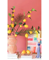 Contre un mur rose corail se trouvent un vase rayé rempli de branches de citron Kunsttak de 104 cm en jaune, un tableau coloré, une pile de livres, un chandelier bleu avec une bougie et un plateau de citrons.