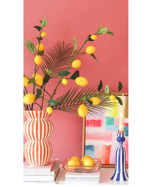 Tegen een koraalroze muur staan een gestreepte vaas gevuld met Kunsttak Lemon-takken van 104 cm in geel, een kleurrijk schilderij, een stapel boeken, een blauwe kandelaar met een kaars en een dienblad met citroenen.