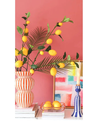Contre un mur rose corail se trouvent un vase rayé rempli de branches de citron Kunsttak de 104 cm en jaune, un tableau coloré, une pile de livres, un chandelier bleu avec une bougie et un plateau de citrons.