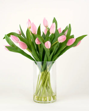 Het Zijden kunstboeket Tulpen in vaas Roze heeft roze tulpen met lange stelen en groene bladeren in een heldere glazen vaas, tegen een effen witte achtergrond.