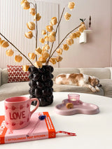 Een gezellige woonkamer met een beige bank, een slapende kat, een sculpturale zwarte vaas met daarin de Kunstbloem Physalis Lampion 90 cm bruin, een roze "LOVE" mok, een boek, een kaars en een koekje op de salontafel.